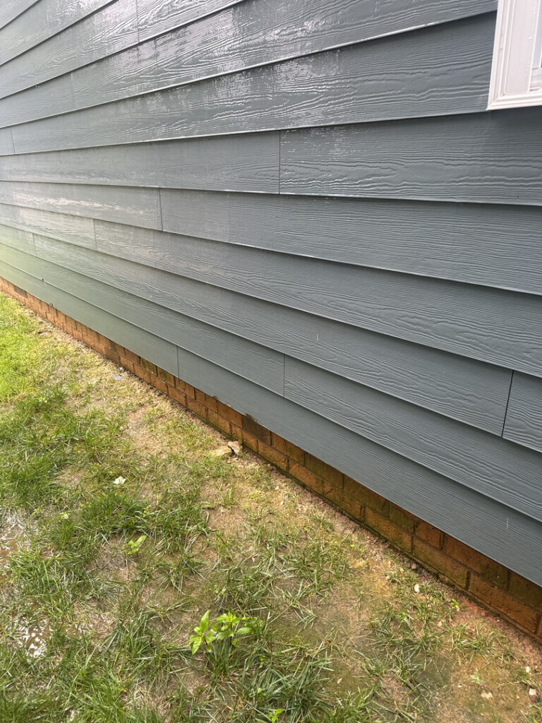 green vinyl siding being cleaned with water exposure on lower sections and wet grass around the base